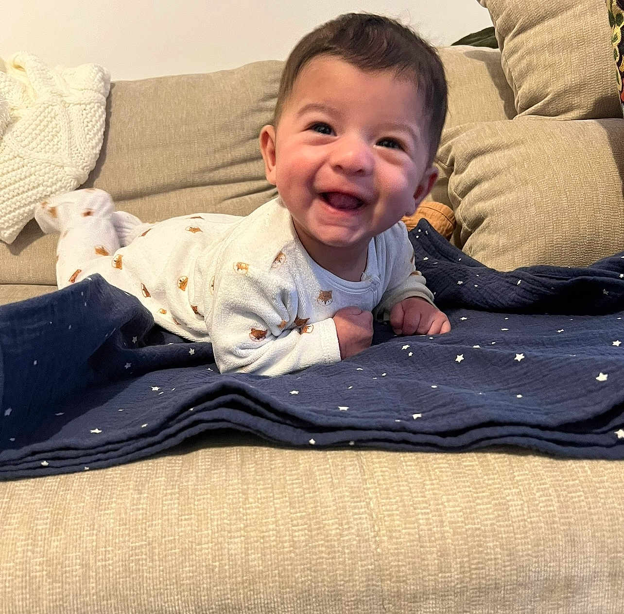 Sohan participe au concours pour gagner de l'argent avec cette photo : baby, smiling, infant, onesie, blanket, couch, indoor, happy, face, child, cute, lying_down, cozy, home, furniture, patterned_clothing, star_pattern, toddler, joyful, portrait
