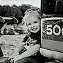 child, smiling, girl, curly_hair, bow, construction_vehicle, black_and_white, outdoor, jeans, tshirt, happy, portrait, sitting, vehicle, metal, rustic, playful, young, casual, cute