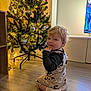 child, christmas_tree, clothing, cute, decorations, face, festive, floor, furniture, holiday, holiday_season, home, indoor, lights, ornaments, person, smile, socks, toddler, wood_floor