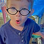 child, boy, glasses, blue_shirt, outdoor, expression, surprised, face, short_hair, standing, chair, table, grass, casual_clothing, daylight, person, young, curious, portrait, funny_face