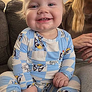 Wade is registered to the contest to win money with this photo: baby, infant, child, pajamas, smile, teeth, couch, living_room, person, blonde_hair, close_up, happy, sitting, patterned_clothing, cartoon_character, furniture, hand, cozy, portrait, home