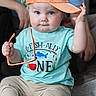 baby, beige_pants, blue_eyes, cap, child, chubby_cheeks, cute, graphic_tee, hand_raised, hardwood_floor, indoor, mesh_cap, neutral_expression, parent_in_background, portrait, sitting, sunglasses, sunglasses_in_hand, t_shirt, toddler