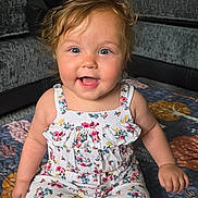 Alina is registered to the contest to win money with this photo: baby, child, smiling, sitting, romper, floral_pattern, curly_hair, blue_eyes, quilt, floor, living_room, sofa, cushion, anklet, barefoot, happy, indoor, person, infant, cute