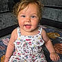 baby, child, smiling, sitting, romper, floral_pattern, curly_hair, blue_eyes, quilt, floor, living_room, sofa, cushion, anklet, barefoot, happy, indoor, person, infant, cute