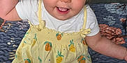 Alina is registered to the contest to win money with this photo: baby, child, smiling, hat, yellow_dress, pineapple_pattern, sitting, indoor, quilt, playful, happy, cute, tongue_out, hand_wave, infant, clothing, footwear_bare, face, young_child, home