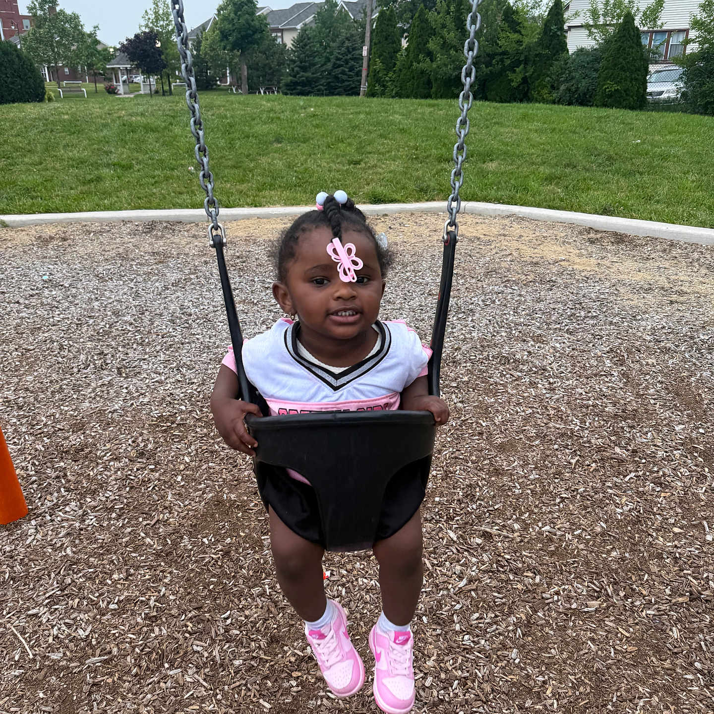 Jersey Gibson is registered to the contest to win money with this photo: toddler, child, swing, playground, grass, trees, house, park, outdoor, sneakers, pink, hair_clip, smiling, person, daylight, wood_chips, play, fun, casual_clothing, happy
