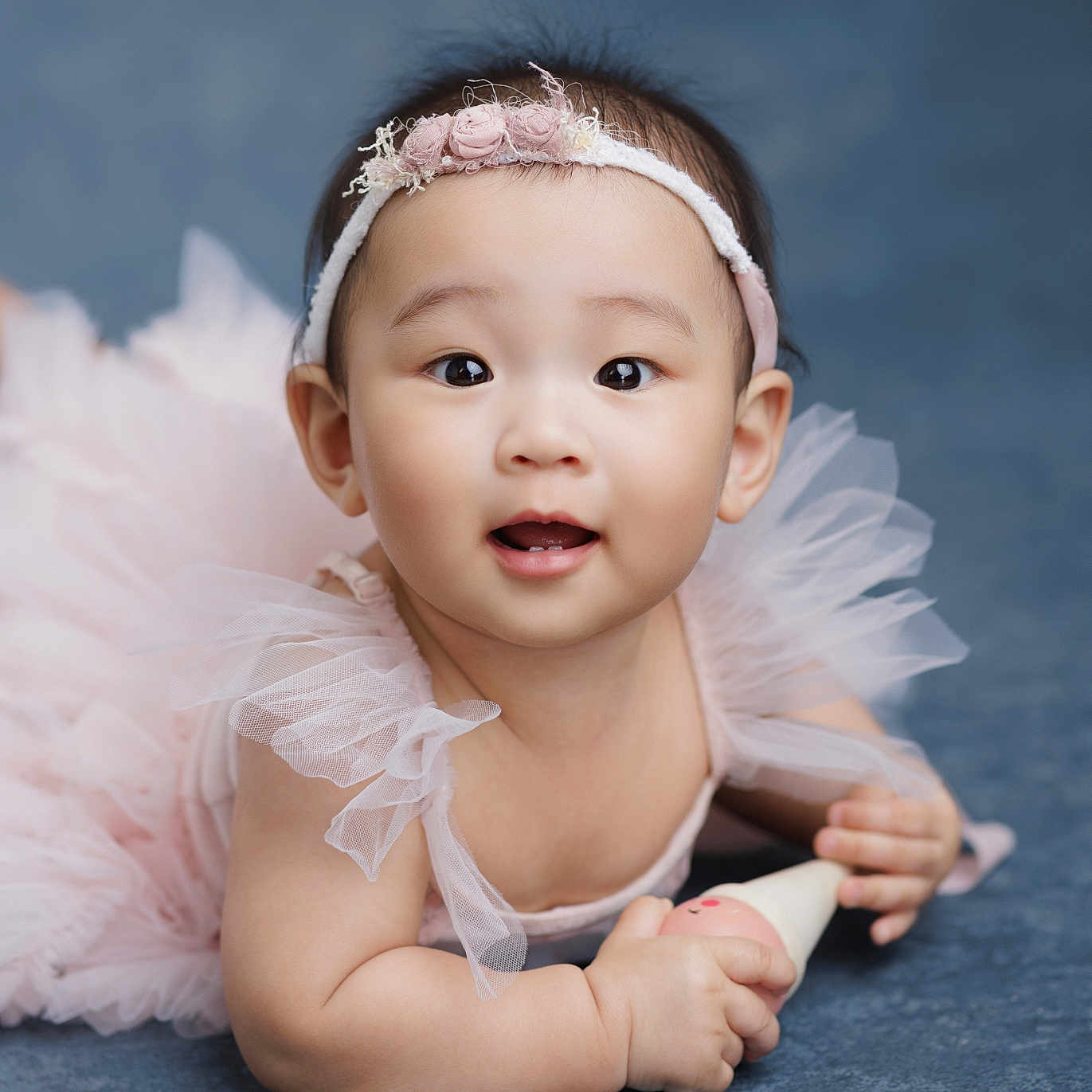 Madelyn joined the competition — help win amazing prizes! baby, child, portrait, smiling, happy, flower_headband, pink_dress, tulle, ruffles, toy, sitting, indoor, studio, blue_background, closeup, infant, fashion, cute, photography, soft_lighting