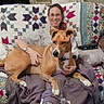 blanket, brown_dog, casual_clothing, comfort, couch, dog, furniture, glasses, happy, home, indoor, living_room, person, pet, quilt, relaxation, remote_control, seated, smile, woman