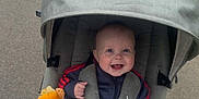 Otis is registered to the contest to win money with this photo: baby, stroller, blanket, plush_toy, smile, happy, outdoor, child, jacket, canopy, pavement, grass, person, cute, infant, walking, hood, comfort, toy, seatbelt