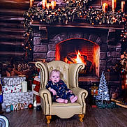 Otis is registered to the contest to win money with this photo: baby, armchair, fireplace, wreath, candles, christmas_decorations, pajamas, wood_floor, gift_boxes, nutcracker, toy, holiday, indoor, cozy, warm_lighting, face, person, floor, decor, sitting
