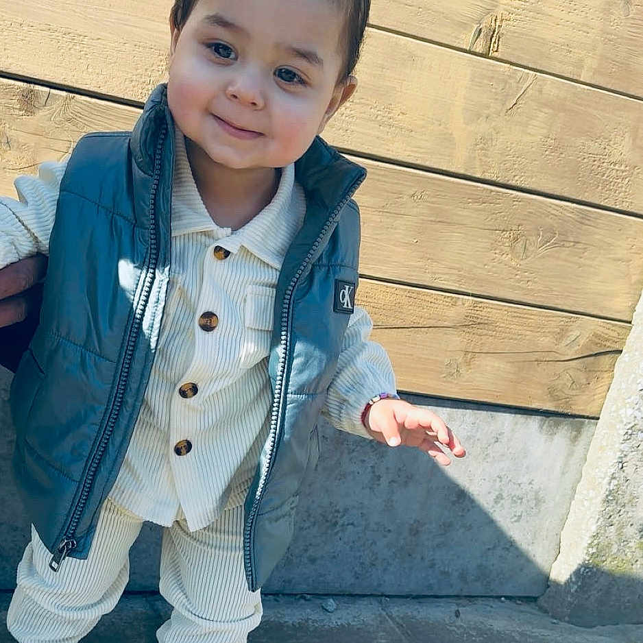 Milan a rejoint le concours — aidez-le/la à gagner de superbes lots ! toddler, child, smiling, outdoor, pavement, wooden_wall, vest, white_shoes, curly_hair, cute, standing, sunlight, shadow, casual_clothing, portrait, young_child, daylight, fashion, person, happy