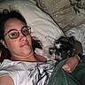 bed, bedroom, blanket, cozy, cuddling, dog, glasses, indoors, long_hair, lying_down, person, pet, pillow, portrait, relaxed, shaggy_fur, small_dog, white_shirt, woman, wristwatch