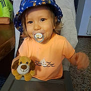 Yann participe au concours pour gagner de l'argent avec cette photo : toddler, child, pacifier, plush_toy, bucket_hat, orange_shirt, high_chair, indoor, smiling, face, person, furniture, table, toy, hair, eyes, mouth, hand, background, home