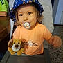 toddler, child, pacifier, plush_toy, bucket_hat, orange_shirt, high_chair, indoor, smiling, face, person, furniture, table, toy, hair, eyes, mouth, hand, background, home