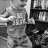 child, toddler, boy, standing, walking, smiling, indoors, portrait, black_and_white, bookshelf, books, clothing, t_shirt, pants, wood_floor, shoes, home, playful, motion, face