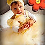 baby, child, dress, yellow, headband, bow, bed, white, cute, portrait, infant, smiling, indoors, soft_lighting, toy, cartoon, flower, hand, footwear, tulle