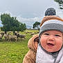 baby, child, smiling, hat, winter_clothing, jacket, bib, outdoor, field, grass, sheep, trees, nature, cloudy_sky, portrait, happy, closeup, person, infant, seasonal