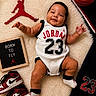 baby, infant, basketball, jordan, sneakers, sportswear, sign, text, smiling, black_hair, white_outfit, red, black, white, sock, rug, toy, child, portrait, indoor