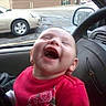 baby, laughing, car_interior, red_clothing, child, happy, person, seat, steering_wheel, window, vehicle, parking_lot, car, joy, smile, head, face, infant, clothing, transport