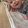 baby, smiling, towel, hand, wooden_surface, plant, shadow, indoor, portrait, child, happy, face, clothing, person, wood, cozy, support, infant, casual, warm_light