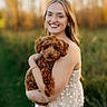 Millie Mae is registered to the contest to win money with this photo: woman, person, puppy, dog, curly_fur, brown_fur, portrait, outdoor, autumn, golden_hour, smiling, hugging, pet, companion, garden, blurred_background, bokeh, floral_top, white_skirt, bracelet