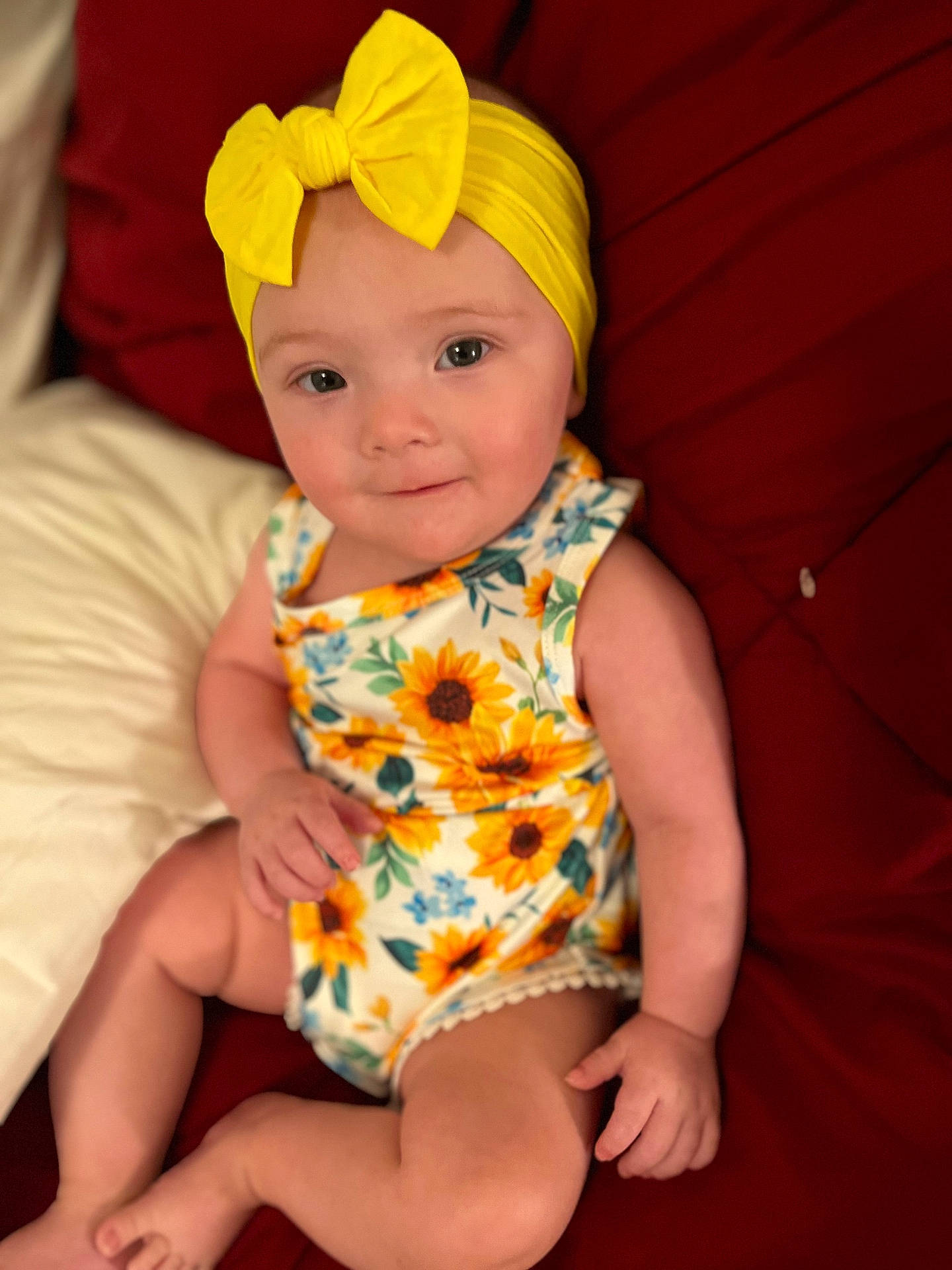 baby, casual, child, closeup, comfortable, cute, floral_outfit, happy, headband, indoors, infant, one_child, person, portrait, red_cushion, sitting, smiling, soft_skin, sunflower, yellow_bow