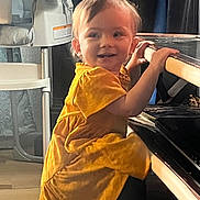 Ellie is registered to the contest to win money with this photo: toddler, child, yellow_dress, piano, indoor, home, smile, curly_hair, furniture, high_chair, wood_floor, window_light, young_child, person, portrait, cute, playing, standing, happy, domestic