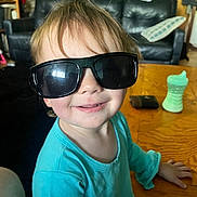 Ellie is registered to the contest to win money with this photo: toddler, child, sunglasses, smile, blue_clothing, table, sippy_cup, wallet, living_room, sofa, pillow, wooden_furniture, face, person, happy, indoors, hair, casual, seat, cute