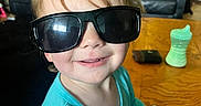 Ellie is registered to the contest to win money with this photo: toddler, child, sunglasses, smile, blue_clothing, table, sippy_cup, wallet, living_room, sofa, pillow, wooden_furniture, face, person, happy, indoors, hair, casual, seat, cute