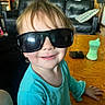 toddler, child, sunglasses, smile, blue_clothing, table, sippy_cup, wallet, living_room, sofa, pillow, wooden_furniture, face, person, happy, indoors, hair, casual, seat, cute