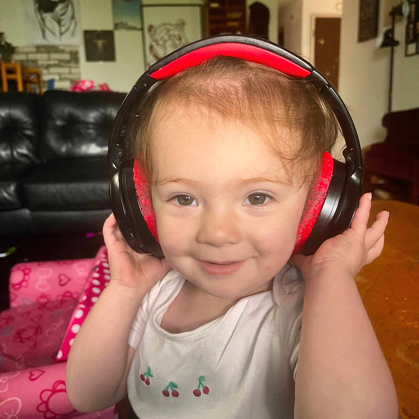 Ellie is registered to the contest to win money with this photo: toddler, child, headphones, smile, indoor, living_room, furniture, couch, pink_chair, wall_art, tiger_drawing, person, cute, happy, casual_clothing, table, head, face, playful, cozy