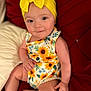 arm, baby, bow, child, clothing, comfort, couch, face, headband, indoors, infant, leg, onesie, pattern, portrait, red_cushion, skin, smile, sunflower, yellow