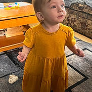 Ellie is registered to the contest to win money with this photo: toddler, child, yellow_dress, standing, indoor, living_room, carpet, coffee_table, toy, furniture, couch, curious, expression, person, home, floor, socks, young_child, portrait, casual