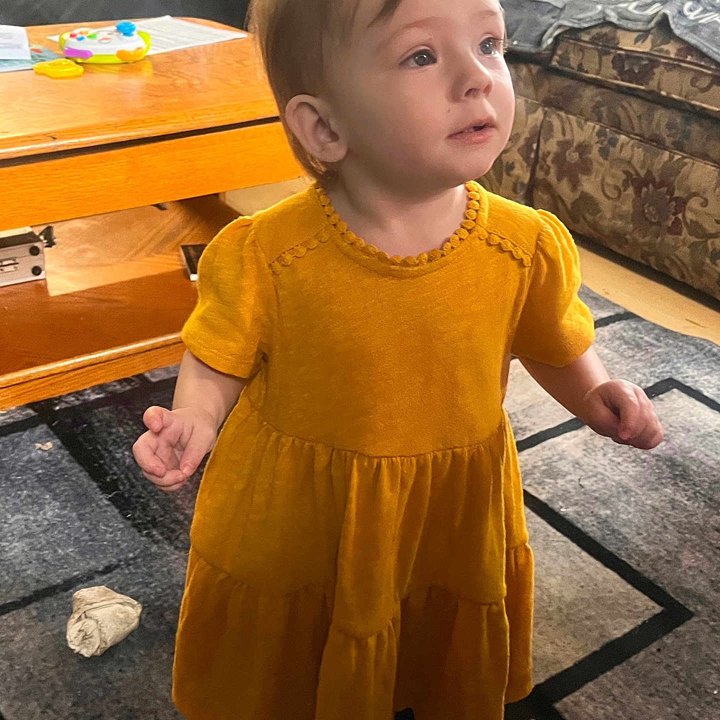 Ellie is registered to the contest to win money with this photo: toddler, child, yellow_dress, standing, indoor, living_room, carpet, coffee_table, toy, furniture, couch, curious, expression, person, home, floor, socks, young_child, portrait, casual