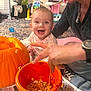 adult_hand, baby, carpet, child, family_activity, fun, happy, indoor, jeans, necklace, newspaper, orange_bucket, pumpkin, pumpkin_carving, pumpkin_seeds, sitting, smiling, toys, watch, woman