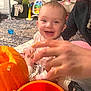 adult, baby, carpet, child, clothing, face, floor, fun, hand, happy, home, indoor, orange_bucket, pattern, person, playful, pumpkin, sitting, smiling, toys