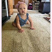 Amelia is registered to the contest to win money with this photo: baby, child, crawling, headband, romper, carpet, living_room, furniture, couch, cabinet, wooden, indoor, toddler, curious, cute, pink, blue, floor, home, person