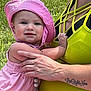 baby, child, pink_hat, pink_outfit, adult_hand, tattoo, yellow_top, grass, outdoor, sunlight, holding, cute, infant, smiling, person, summer, warm, skin, portrait, nature