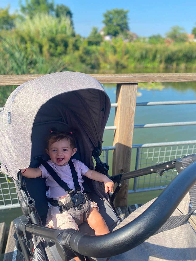 Martina participe au concours pour gagner de l'argent avec cette photo : baby, stroller, outdoor, smiling, child, sunlight, wooden_deck, water, nature, greenery, happy, safety_harness, infant, summer, park, person, playful, cute, daylight, relaxing