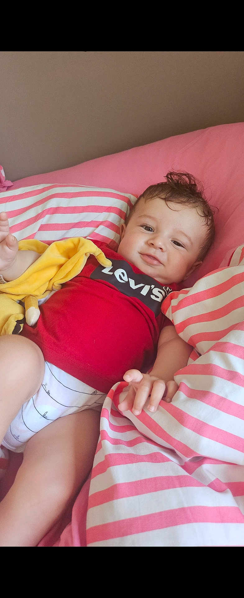Reda participe au concours pour gagner de l'argent avec cette photo : baby, child, infant, red_shirt, levi's, yellow_toy, striped_bedding, pink_bedding, white_bedding, bed, relaxed, smiling, indoors, cute, person, portrait, hand, face, lying_down, soft_toy