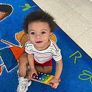 Alexander is registered to the contest to win money with this photo: toddler, child, toy, xylophone, colorful, rug, floor, indoor, smiling, happy, curly_hair, striped_shirt, red_shorts, white_sneakers, playtime, music, bright, eyes, looking_up, young_child