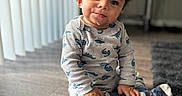 Alexander joined the competition — help win amazing prizes! toddler, child, dinosaur_pajamas, sitting, wooden_floor, crumbs, food, indoors, natural_light, curly_hair, cozy, cute, expression, face, socks, carpet, home, window_blinds, flooring, person