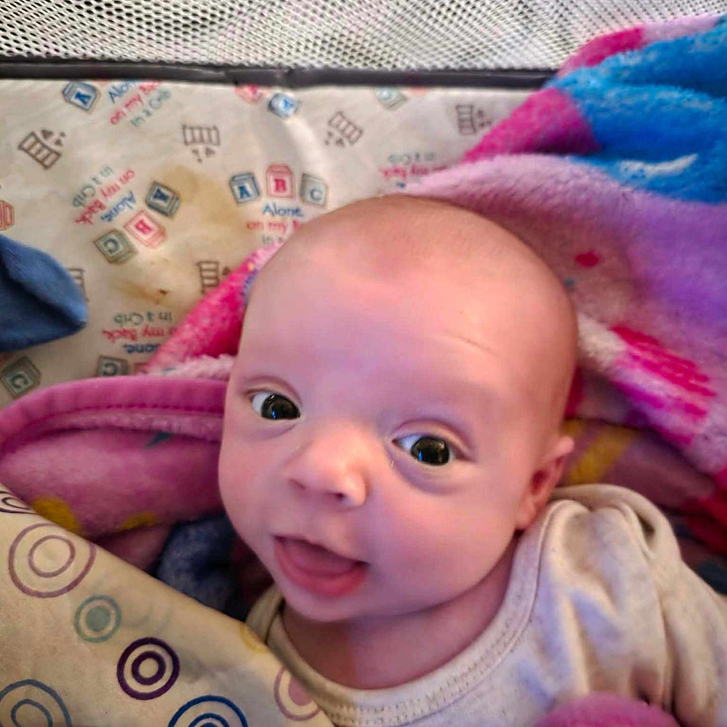 Ezekiel is registered to the contest to win money with this photo: baby, infant, blanket, playpen, face, wide_eyes, curious, expression, cozy, colorful, pattern, clothing, child, indoors, portrait, cute, smile, head, skin, young