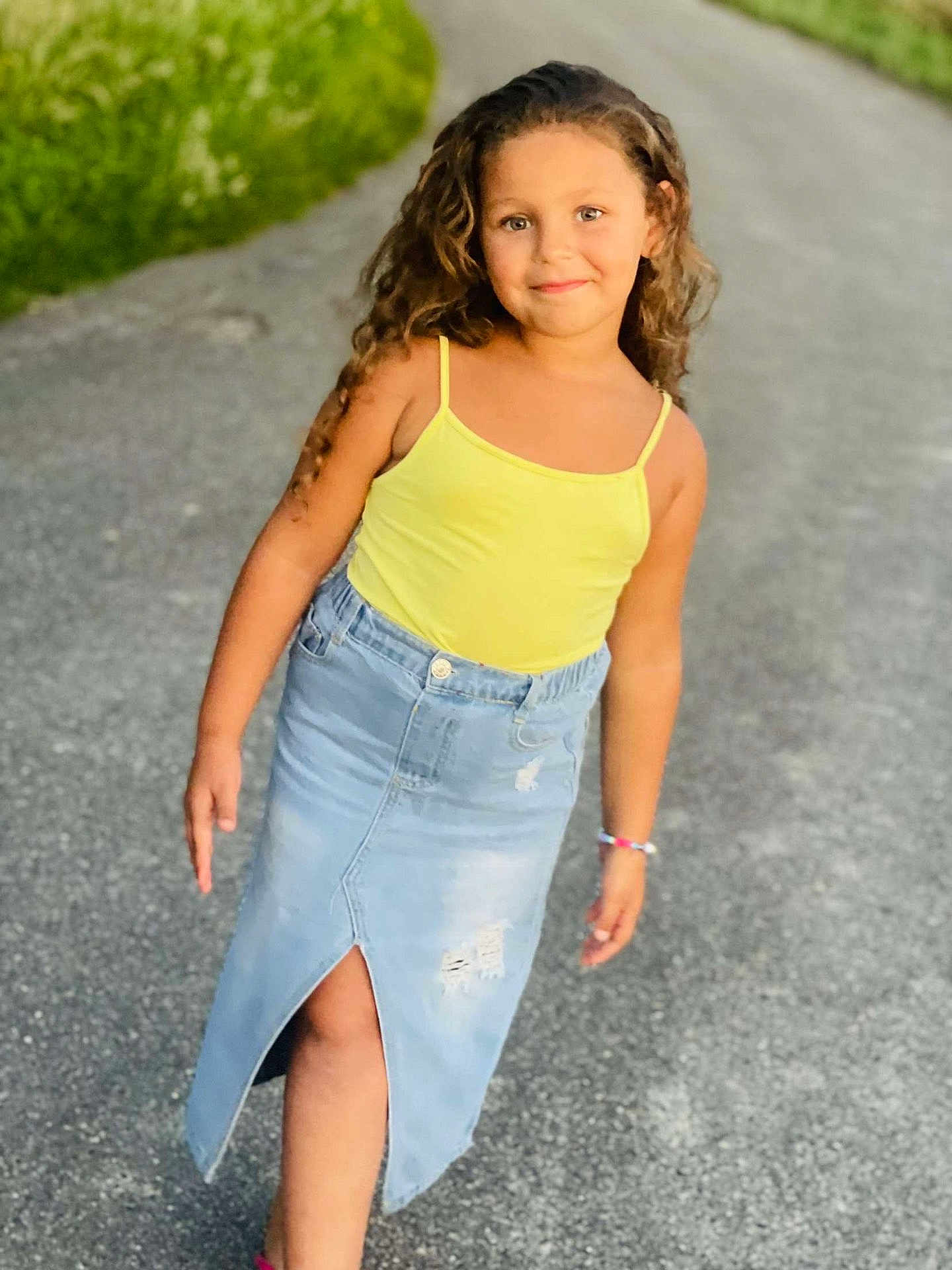 Sarah participe au concours pour gagner de l'argent avec cette photo : child, girl, curly_hair, yellow_top, denim_skirt, sidewalk, outdoor, greenery, smile, bracelet, casual, fashion, portrait, person, happy, young, walking, summer, daylight, road