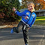 child, boy, smiling, blue_jacket, toy_car, outdoor, fall, autumn_leaves, tree, house, pavement, playful_pose, sneakers, grass, curb, daylight, happy, casual_clothing, young, person