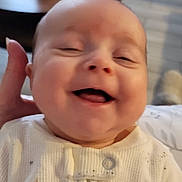 Kaylee is registered to the contest to win money with this photo: baby, smiling, infant, white_sleeper, hand, indoor, living_room, candle, wooden_table, blurred_background, happy, cute, child, person, face, sitting, cozy, warm_lighting, home, expression