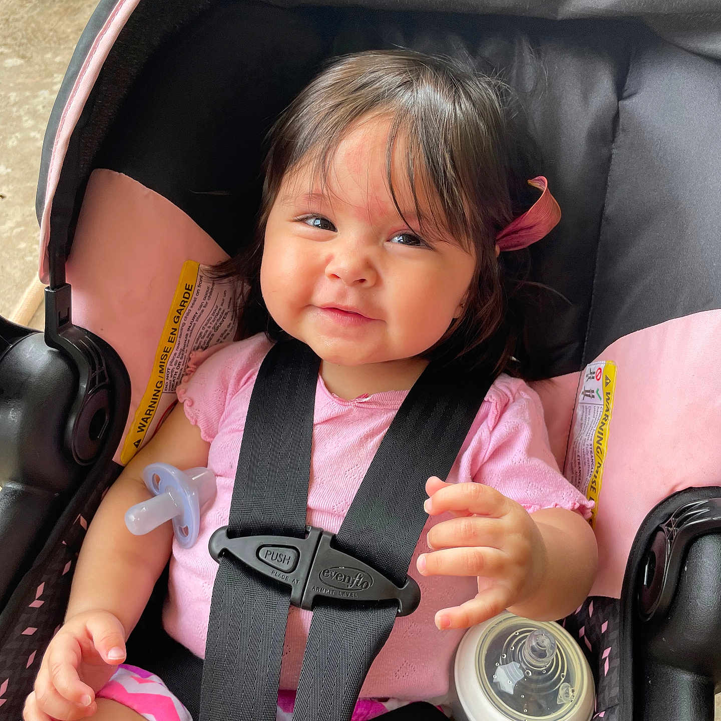 Xylenah is registered to the contest to win money with this photo: baby, bottle, car_seat, child, child_seat, clothing, cute, face, hair_clip, hand, indoor, infant, pacifier, person, pink_clothing, safety_harness, seatbelt, sitting, smile, transport