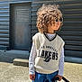 blue_jeans, casual_clothing, child, concrete, curly_hair, daylight, door, fashion, los_angeles_lakers, metal_wall, outdoor, person, shadow, side_profile, standing, stroller, sunglasses, sunlight, sweater_vest, urban