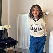 Isaïah a rejoint le concours — aidez-le/la à gagner de superbes lots ! child, curly_hair, smiling, indoor, sweater_vest, blue_pants, hands_in_pockets, radiator, vase, white_roses, table, wooden_floor, casual_clothing, portrait, person, happy, standing, home_interior, decor, fashion
