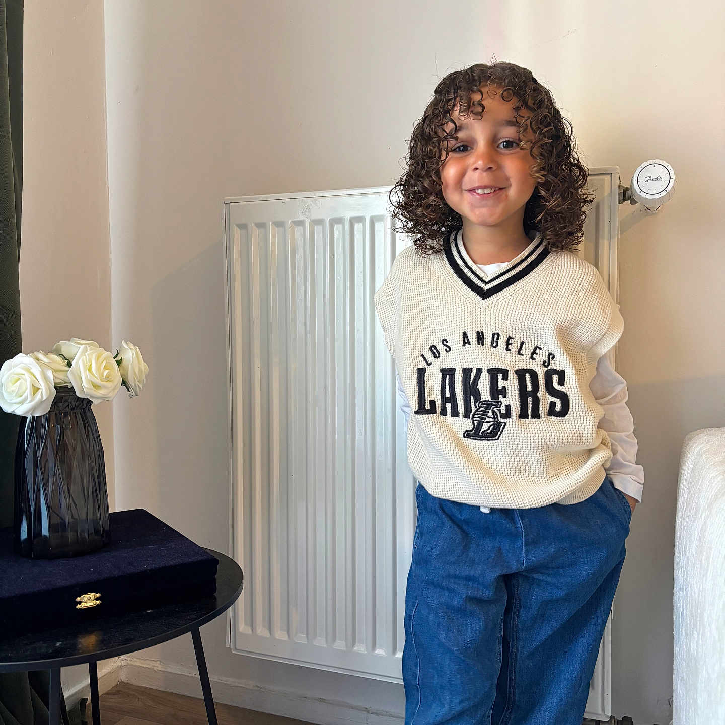 Isaïah a rejoint le concours — aidez-le/la à gagner de superbes lots ! blue_pants, casual_clothing, child, curly_hair, decor, fashion, hands_in_pockets, happy, home_interior, indoor, person, portrait, radiator, smiling, standing, sweater_vest, table, vase, white_roses, wooden_floor